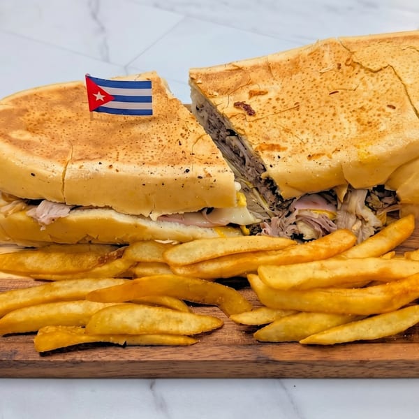 Cuban sandwich cut in half with a Cuban flag toothpick, served with golden french fries on a wooden board.